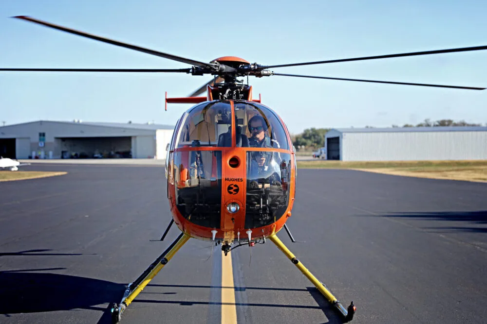 A Hughes 369HS helicopter with an orange front sits on an airport tarmac, viewed from the front.