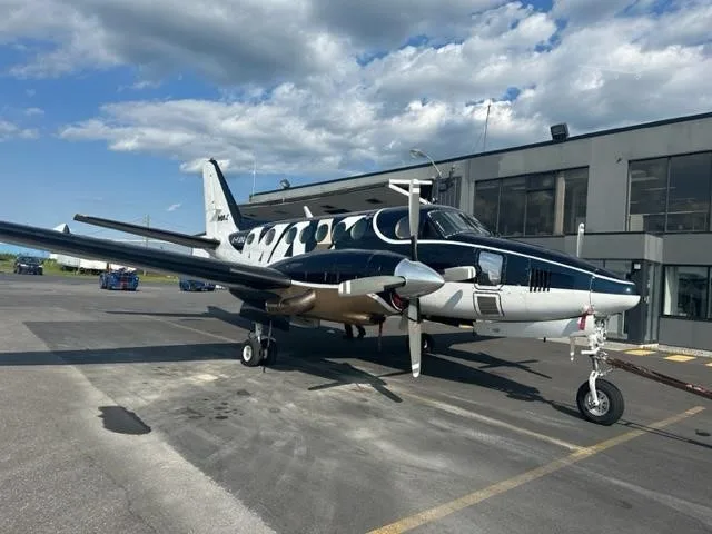 A twin-engine turboprop aircraft with a dark blue, white, and gold paint scheme, parked on an airport apron next to a building.