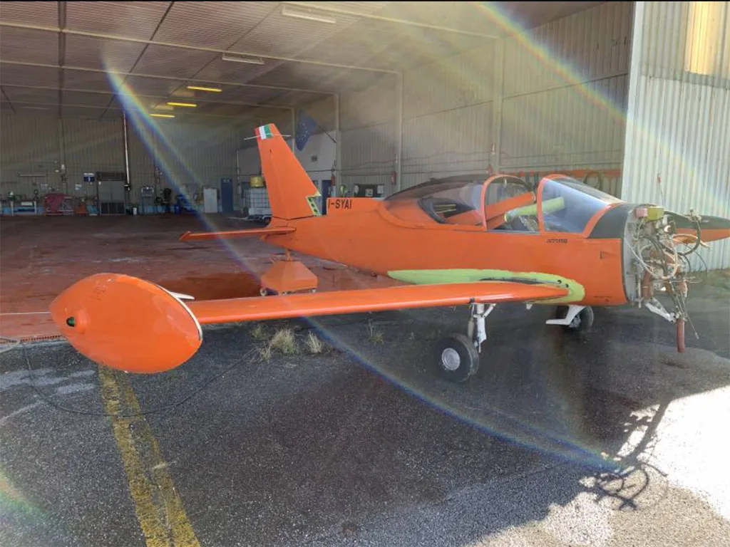 An orange single-engine aircraft with its nose cowling removed, parked inside a hangar with sunlight streaming in.