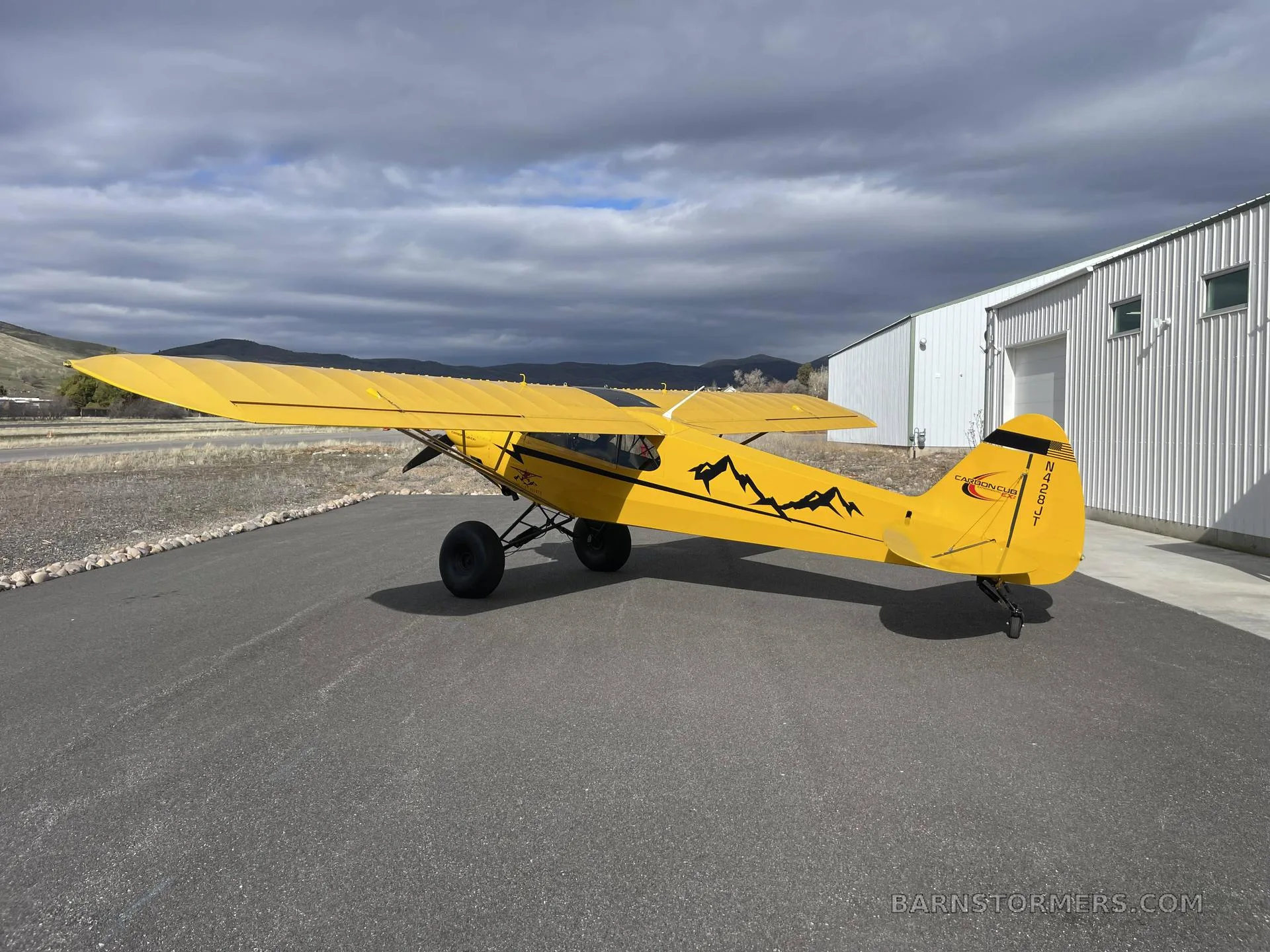 A three-quarter rear view of a yellow CubCrafters Carbon Cub EX-2 with mountain decals and tail number N429ST, parked next to a white hangar.