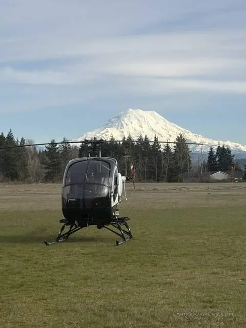 A black helicopter sits on a grassy field with a snow-capped mountain and trees in the background under a partly cloudy sky.