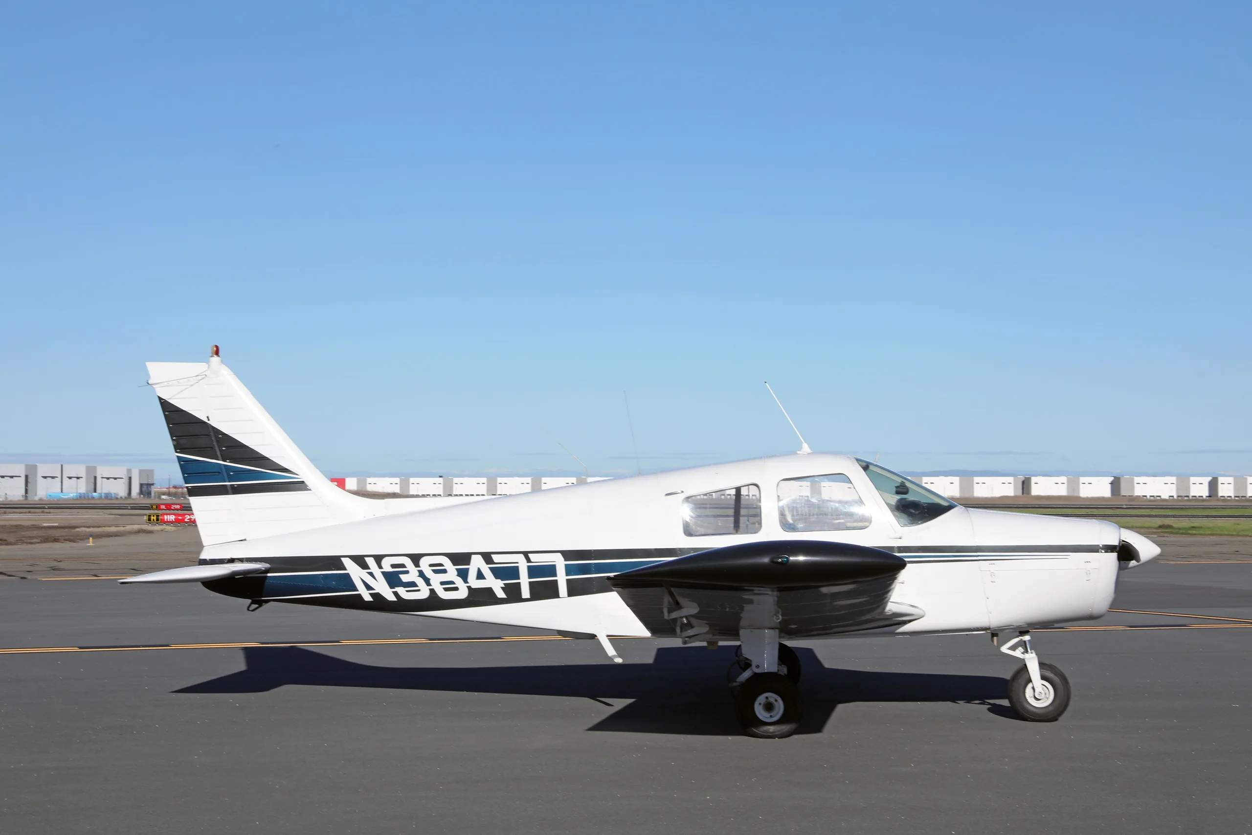 A white Piper Cherokee aircraft with blue and black stripes, registration N38477, is parked on an airfield.