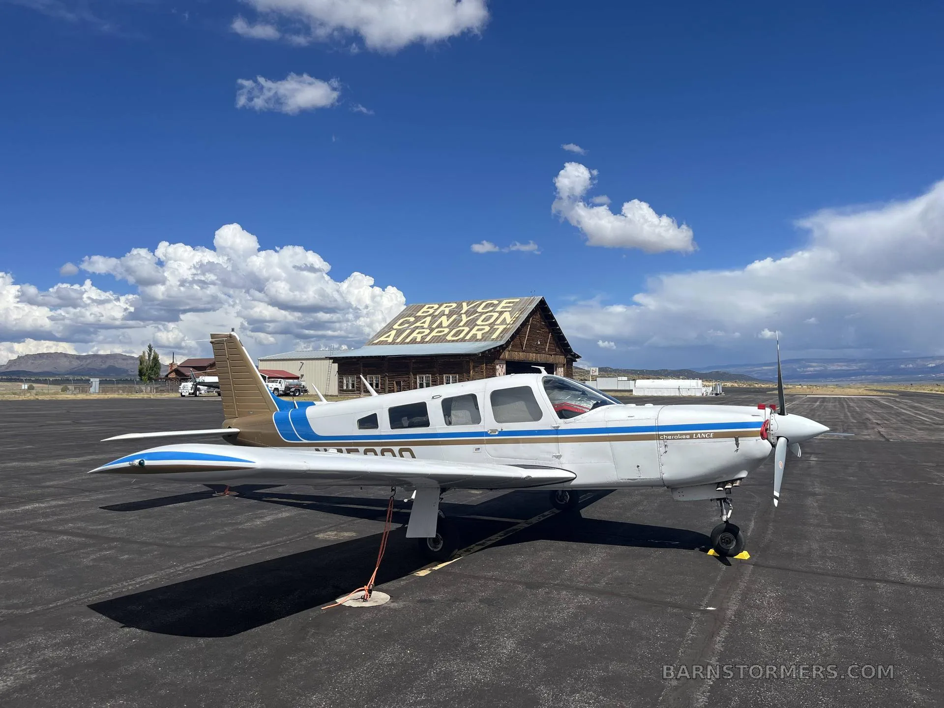 A white and blue Piper Cherokee Lance (N75398) parked on an asphalt ramp in front of the "Bryce Canyon Airport" building under a partly cloudy sky.