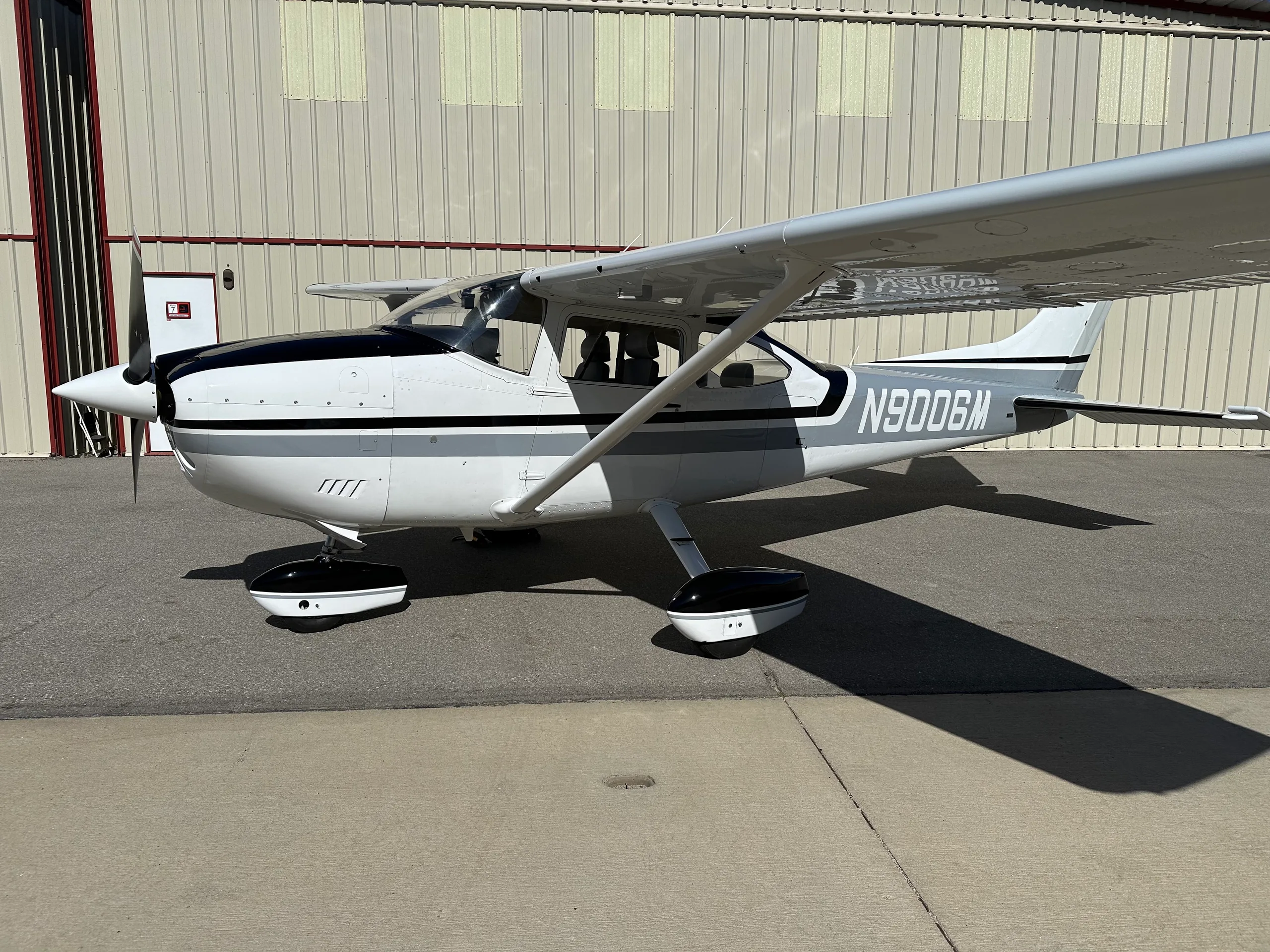 A white and grey Cessna 182 aircraft with registration N9006M parked on an asphalt ramp outside a hangar.
