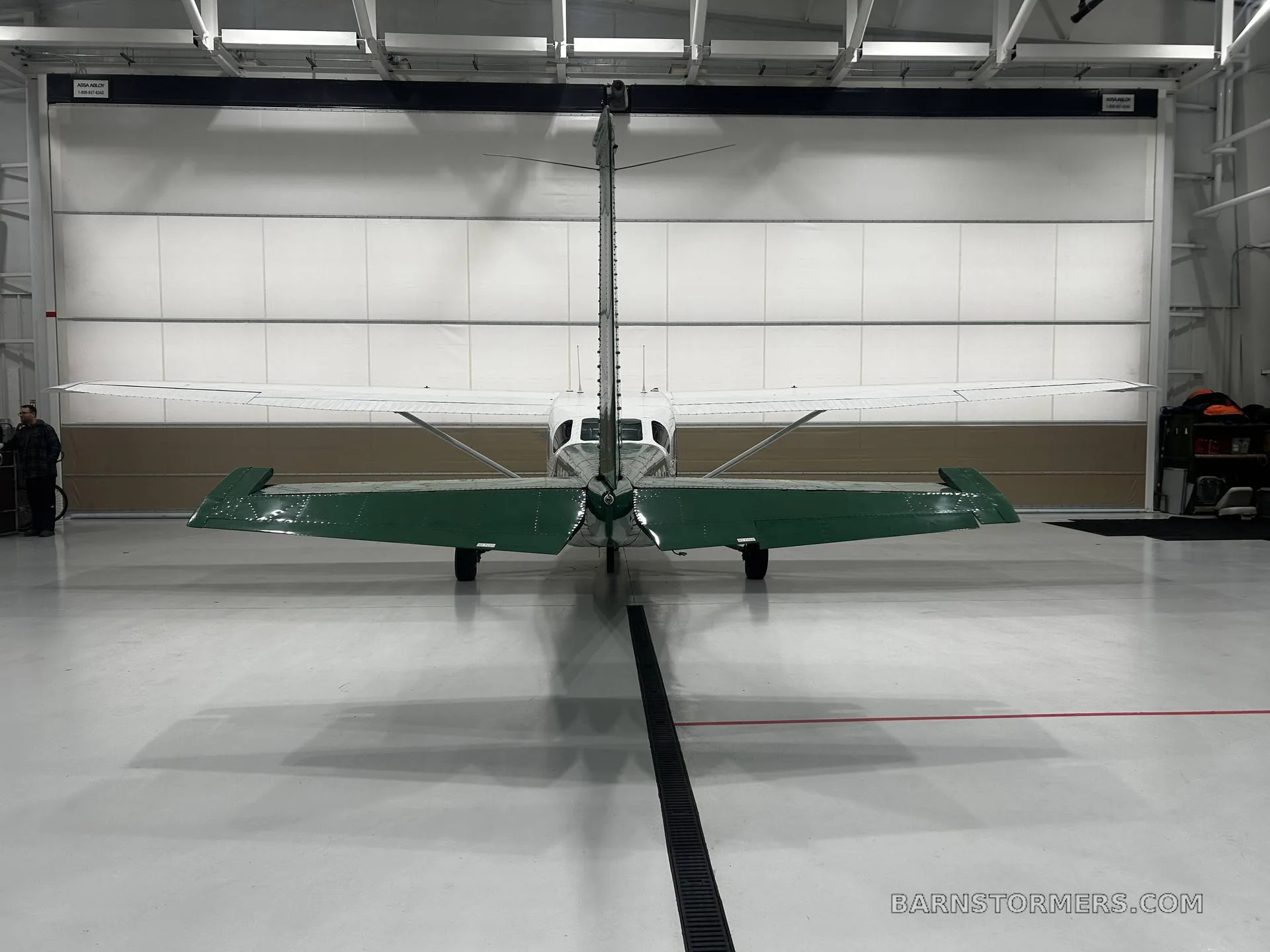 A white and green Cessna 207 Stationair is parked tail-on in a hangar, showing its high wing and tail.