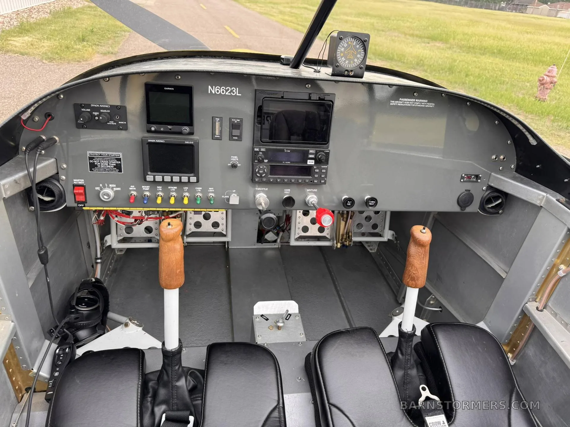 The cockpit of a yellow aircraft features a grey instrument panel with multiple screens, switches, and two wooden control sticks.