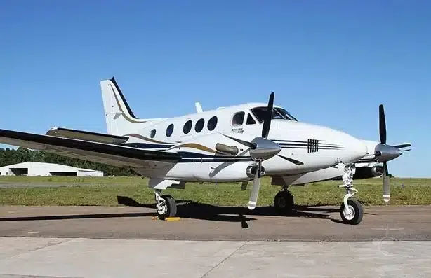 A white twin-turboprop Beechcraft King Air aircraft with gold and black stripes is parked on an airfield under a clear blue sky.