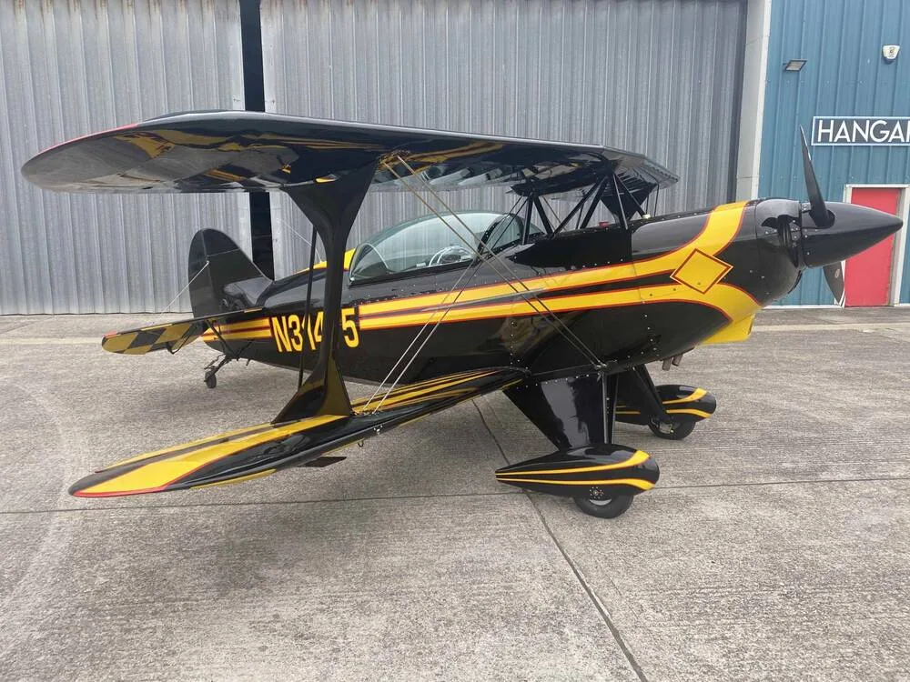 A black and yellow biplane, N31475, is parked on an airfield next to a hangar.