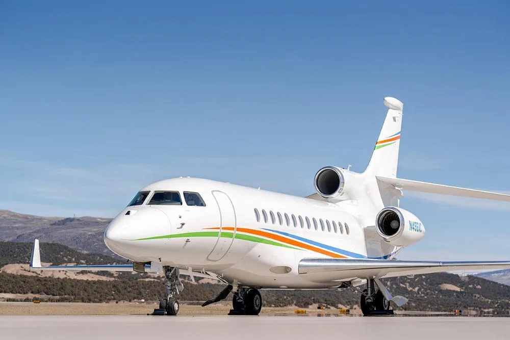 A front-side view of the white Dassault Falcon 900EX, registration N496AC, with colorful stripes, parked on an airfield.