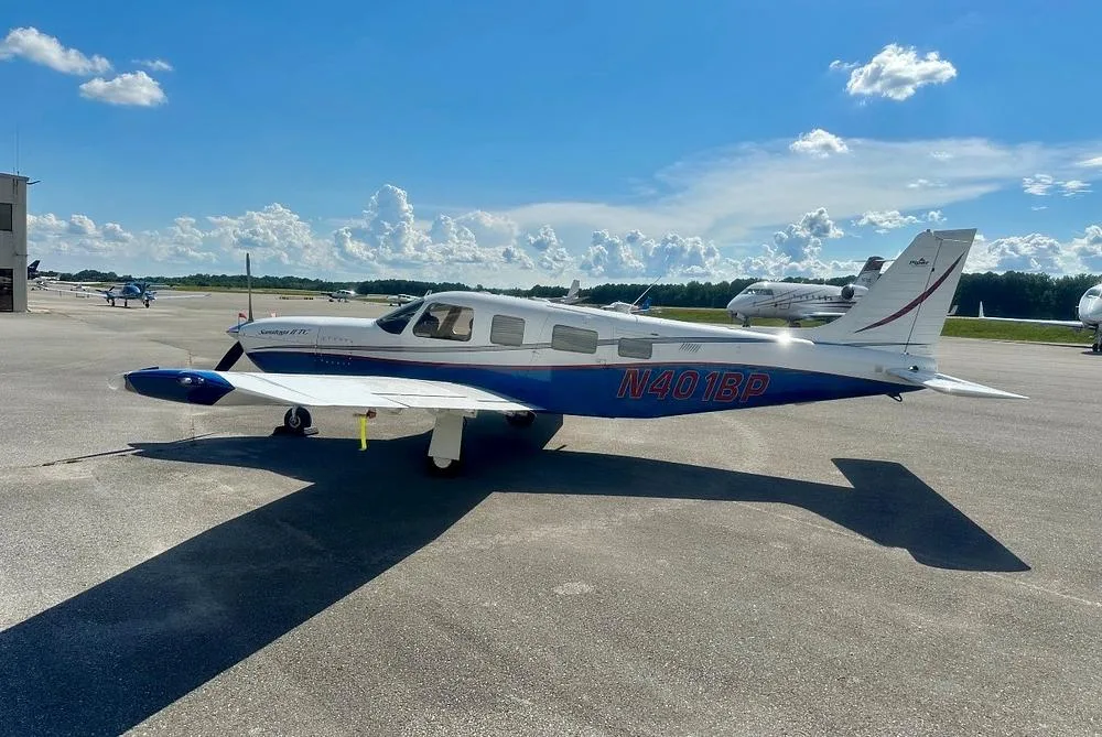 A side view of the Piper Saratoga II TC aircraft, N401BP, in white and blue, parked on an airport tarmac with other aircraft in the background.
