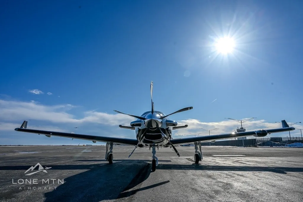 A low-angle front view of a TBM 940 aircraft parked on an asphalt ramp, with the sun brightly shining overhead.