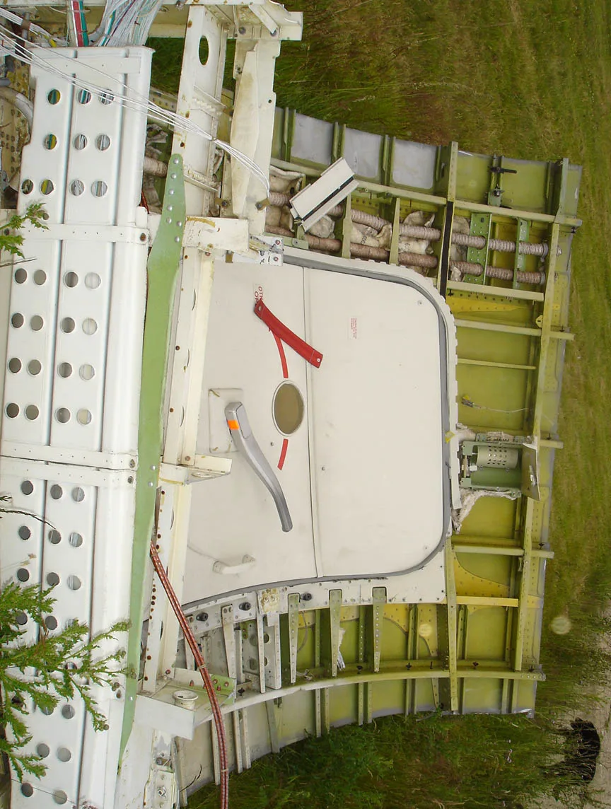 A side view of a disassembled aircraft fuselage section with an emergency exit door and exposed framework.
