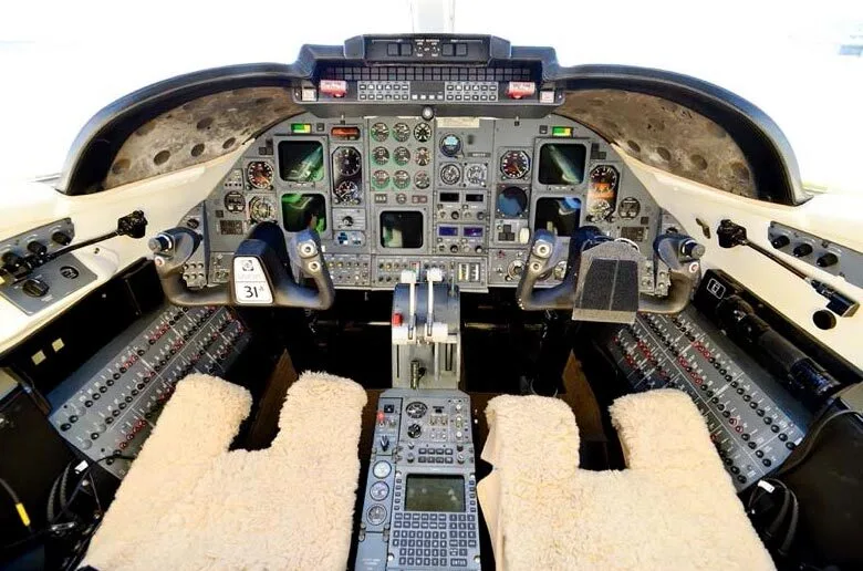 The cockpit of an aircraft showing the instrument panel with various gauges, screens, and controls, featuring sheepskin seat covers.