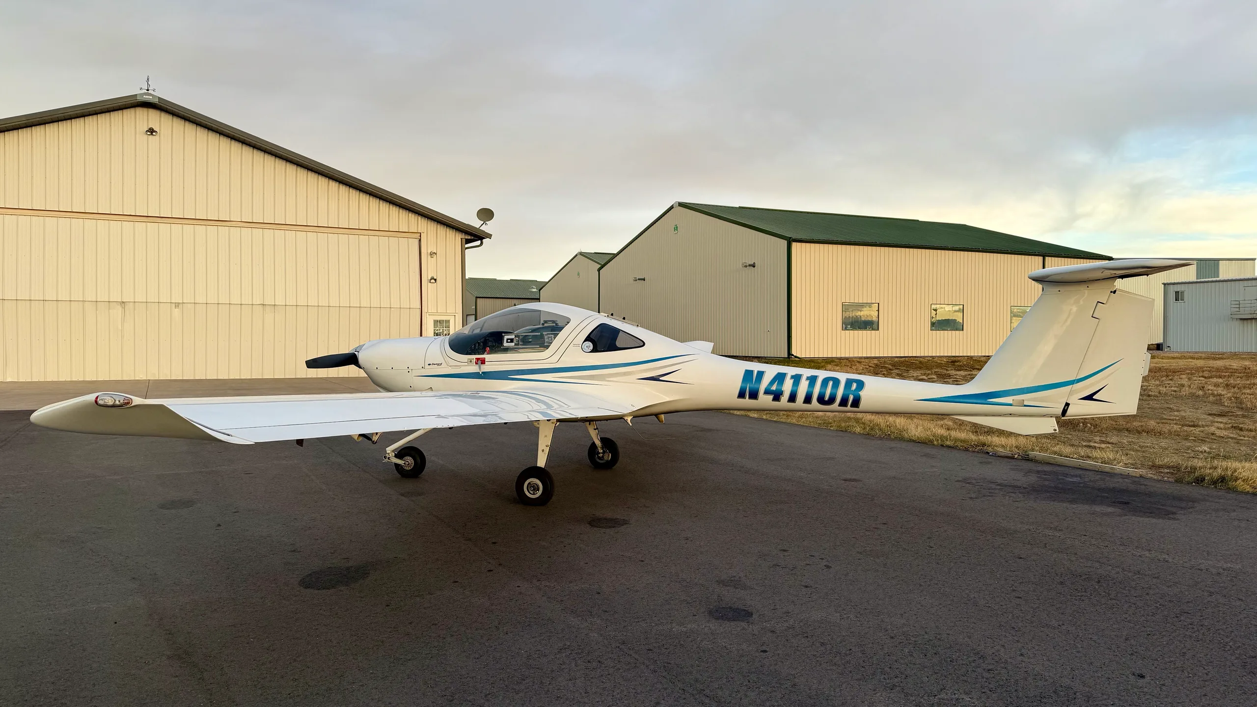 A white Diamond DA20-C1 Eclipse aircraft with blue stripes, registration N4110R, parked on an airfield in front of hangars.
