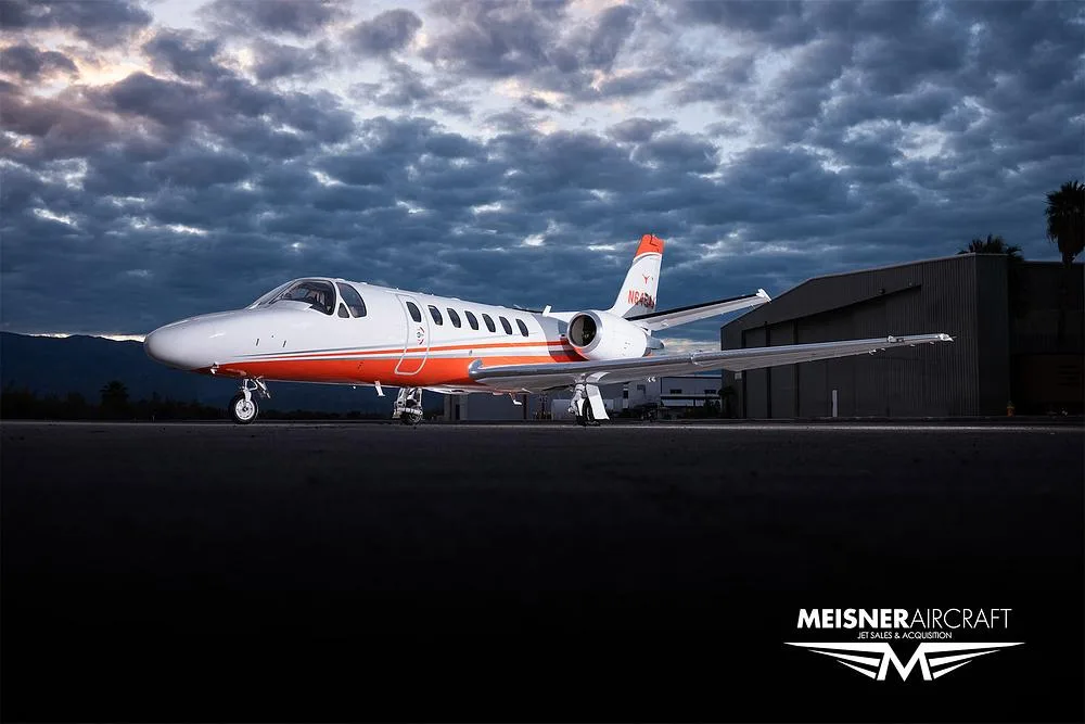 A white and orange Cessna Citation Excel (N648AJ) parked on an airfield at dusk with dramatic clouds.