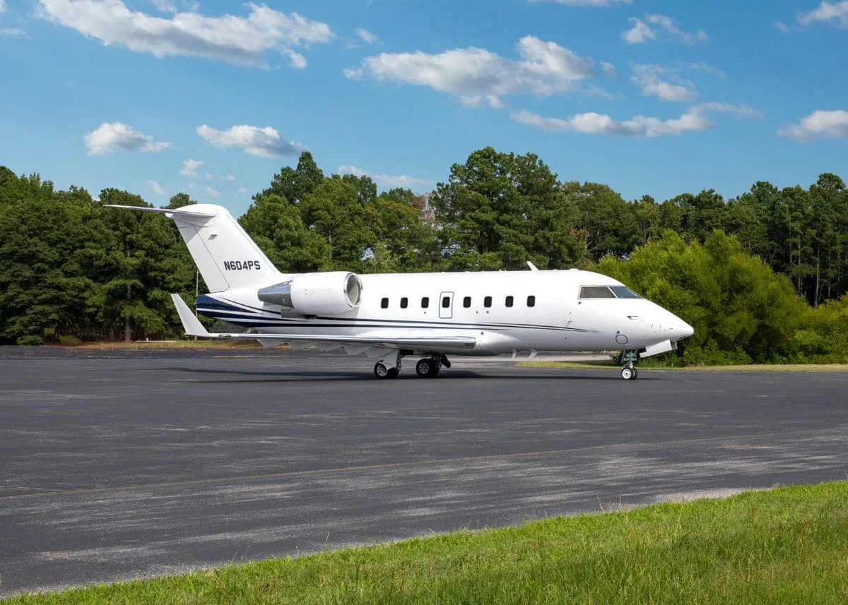 A white Bombardier Challenger 604 business jet (N604PS) with blue stripes on the tail, parked on an asphalt tarmac with green grass and trees in the background.
