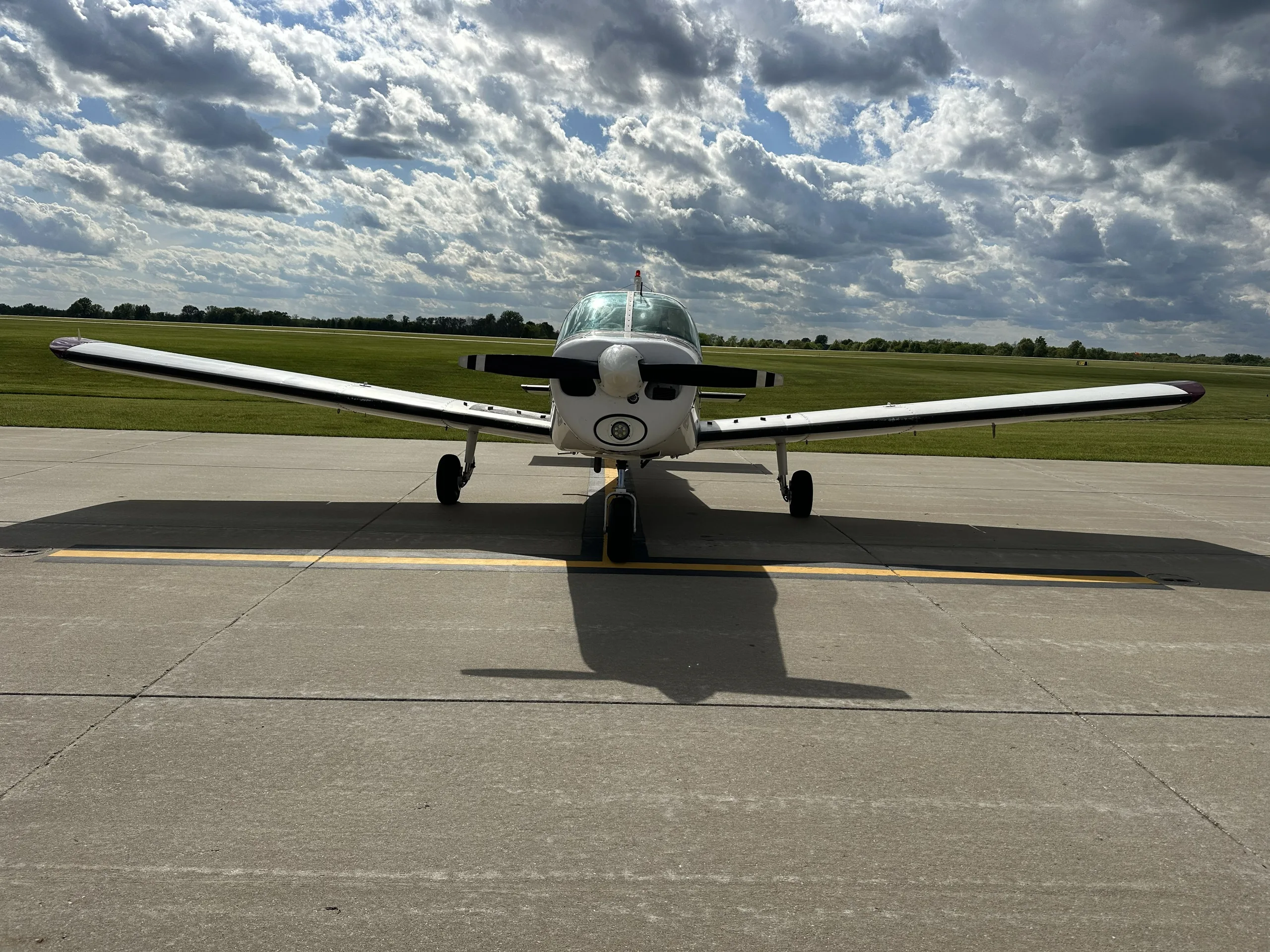A head-on view of a white and maroon single-engine aircraft, likely a Piper Cherokee, parked on an asphalt ramp under a partly cloudy sky.