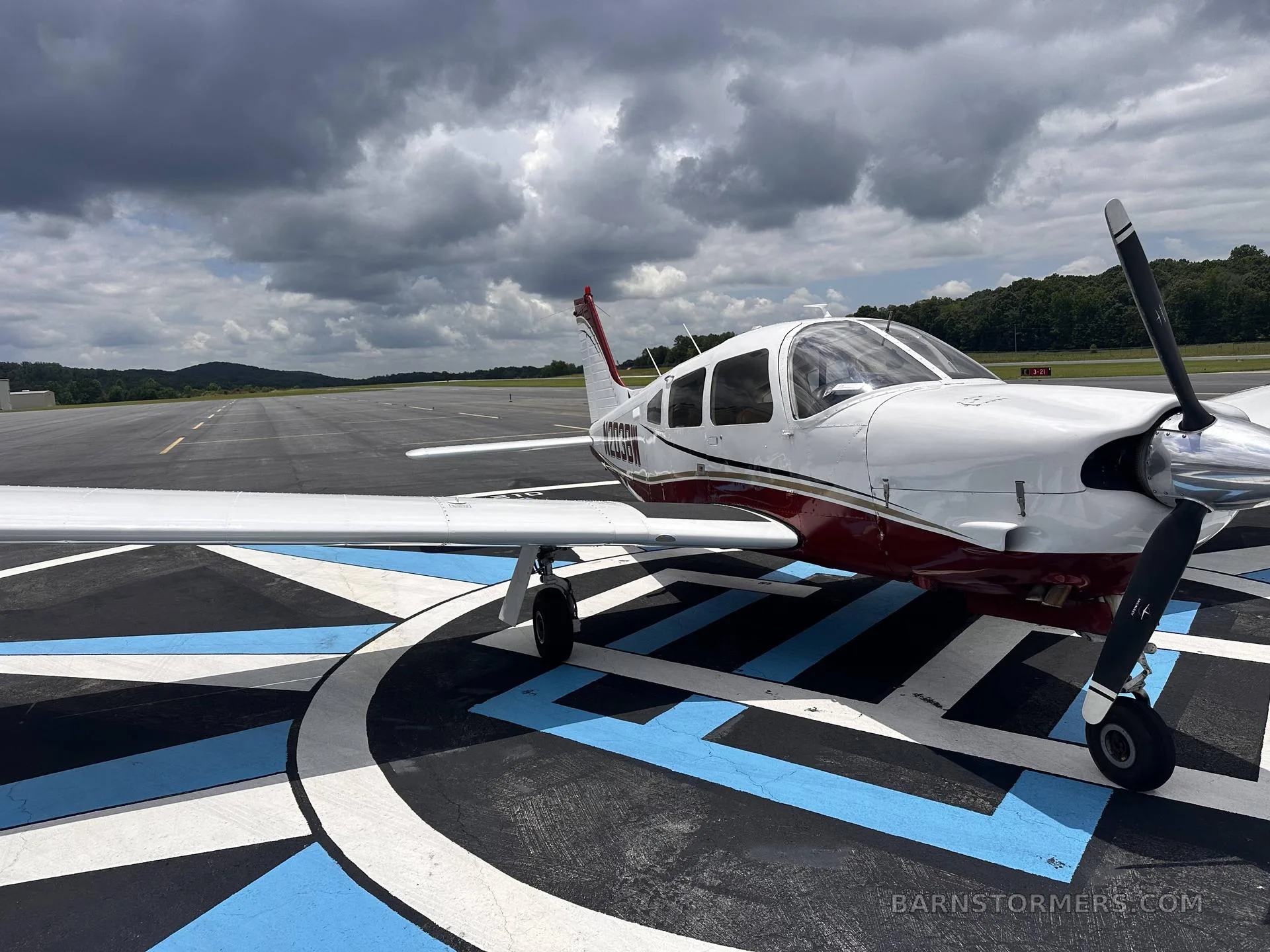 A white and maroon single-engine propeller aircraft (N203BW) is parked on a blue and white patterned tarmac with a runway in the background under a cloudy sky.