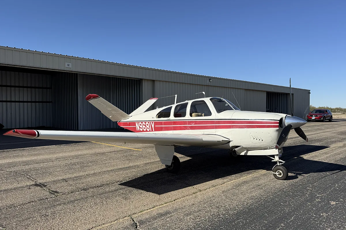 A white single-engine propeller aircraft with red stripes is parked on an asphalt ramp in front of a hangar under a clear blue sky.