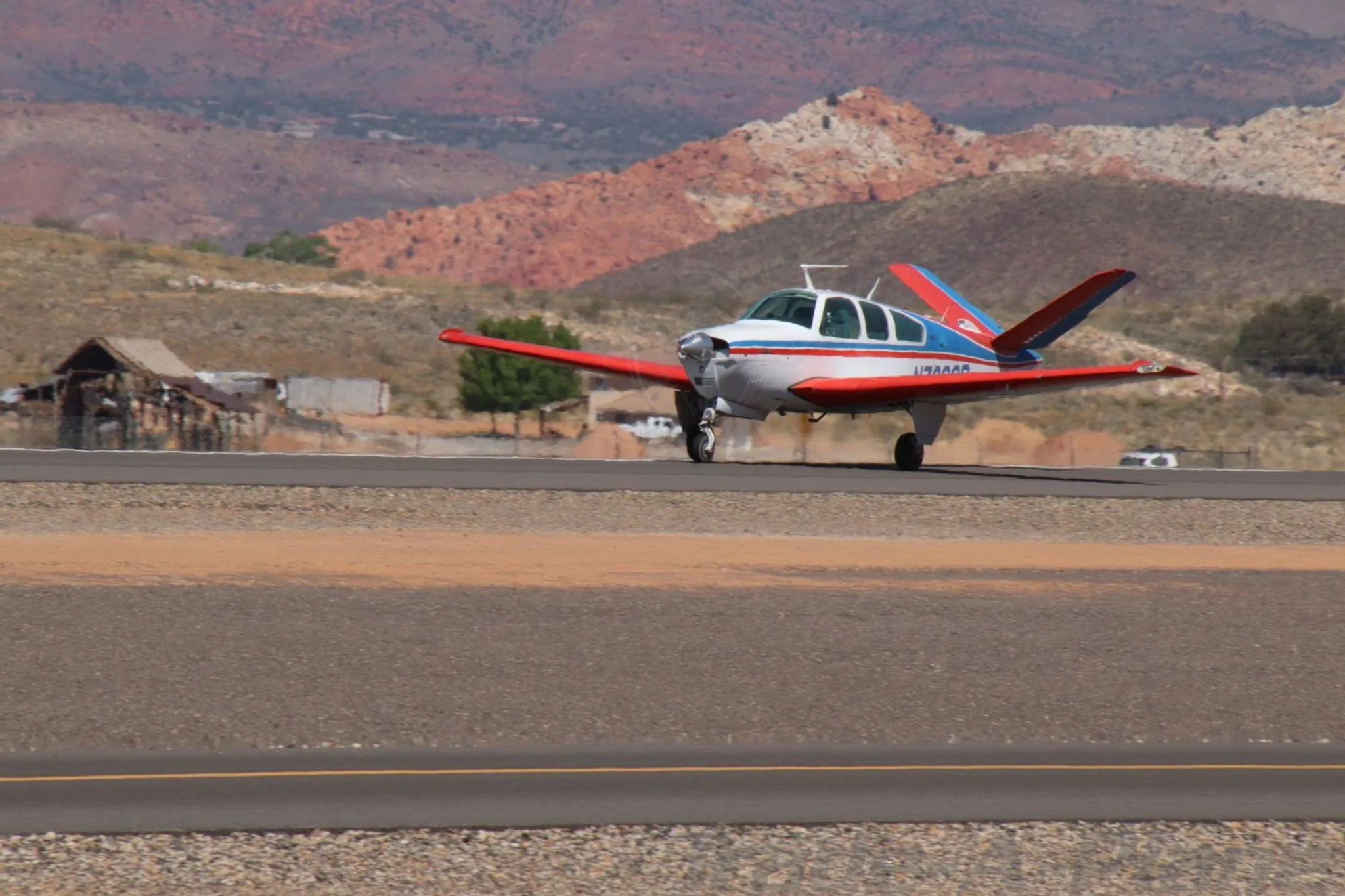A white, red, and blue Beechcraft Bonanza N7208R is on a runway with red-hued mountains in the background.