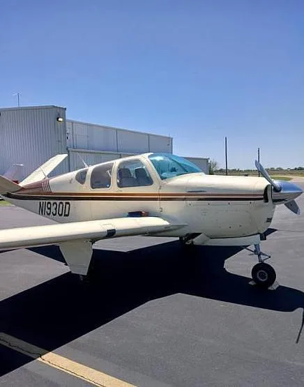 A white and brown single-engine aircraft, N1930D, parked on an asphalt tarmac with a hangar in the background.