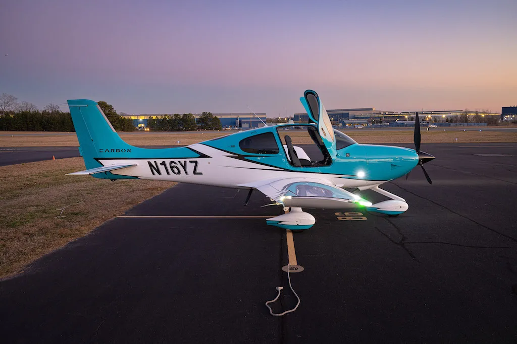 A side view of a turquoise and white Cirrus SR22 aircraft with its doors open, parked on an airfield at dusk.