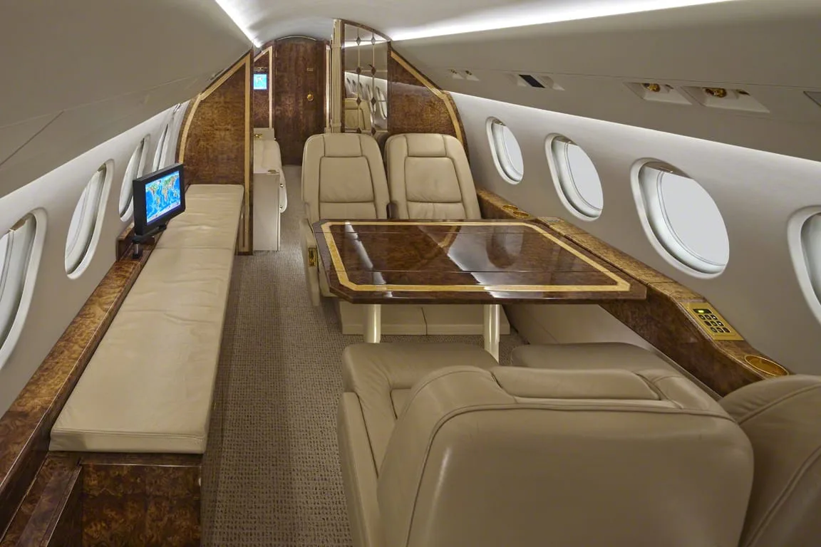 The luxurious interior of a private jet cabin with beige leather seats, a polished wood table, and a TV screen.