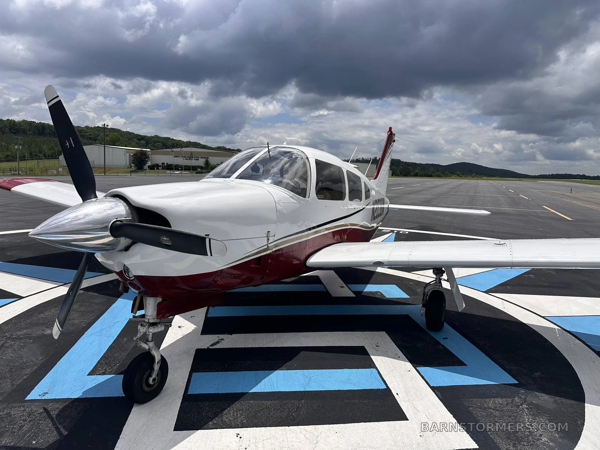 A white and maroon single-engine propeller aircraft (N203BW) is parked at an angle on a blue and white patterned tarmac under a cloudy sky.