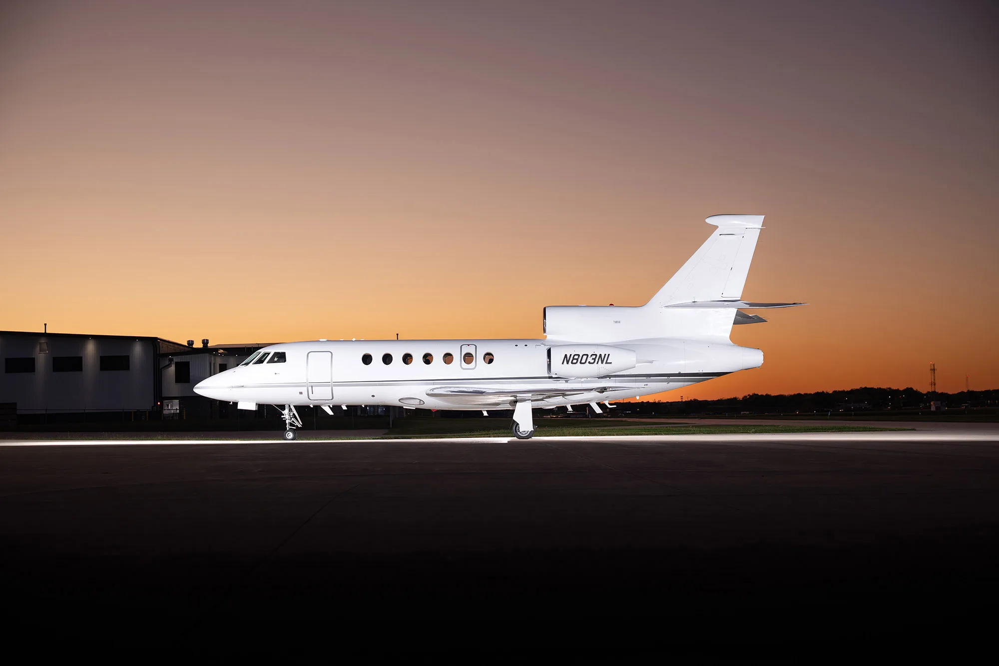A white Dassault Falcon 900EX business jet (N803NL) parked on an airfield at sunset, side view.