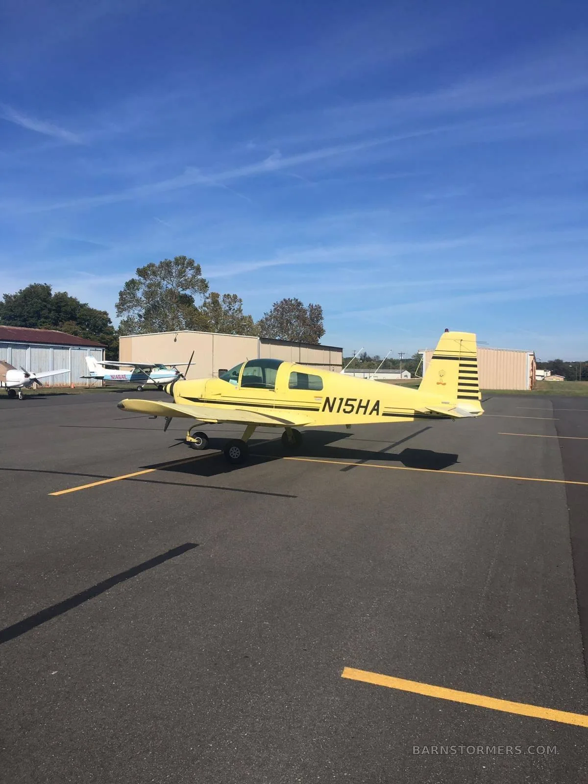 A bright yellow Grumman American AA-1 light aircraft with black stripes and registration N15HA parked on an airport tarmac under a clear blue sky.