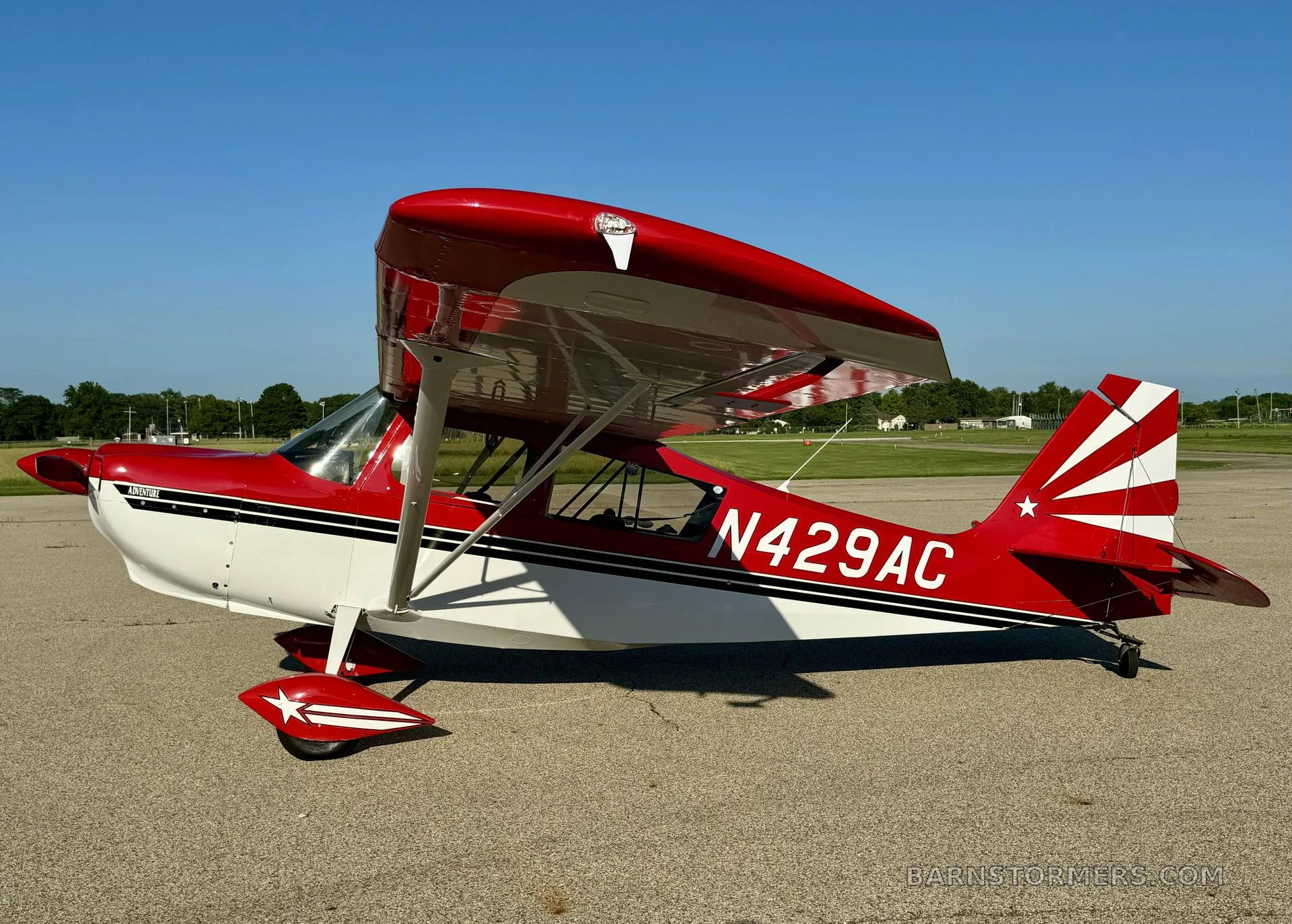 A red and white Adventure aircraft, N429AC, with a star on the tail and wheel fairings, parked on an airfield under a clear blue sky.