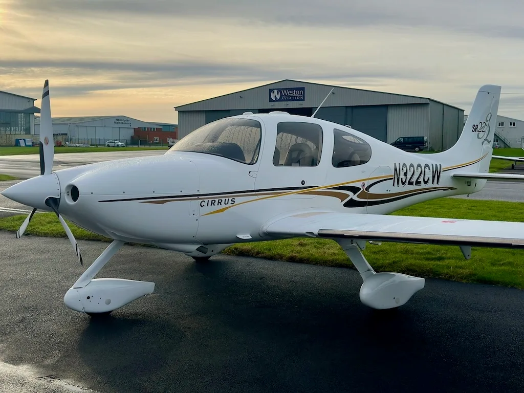 A front-side view of a white Cirrus SR22 aircraft with N322CW registration, parked on a tarmac with hangars under a cloudy sky.