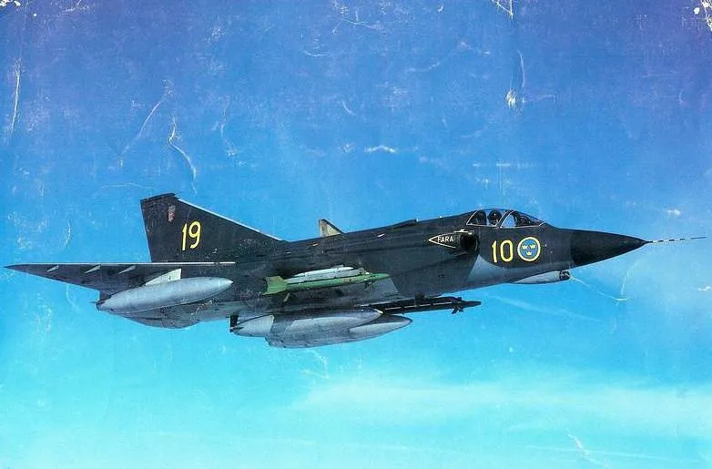 A dark military Saab J35 Draken fighter jet with yellow markings flies against a blue sky, carrying missiles and fuel tanks.
