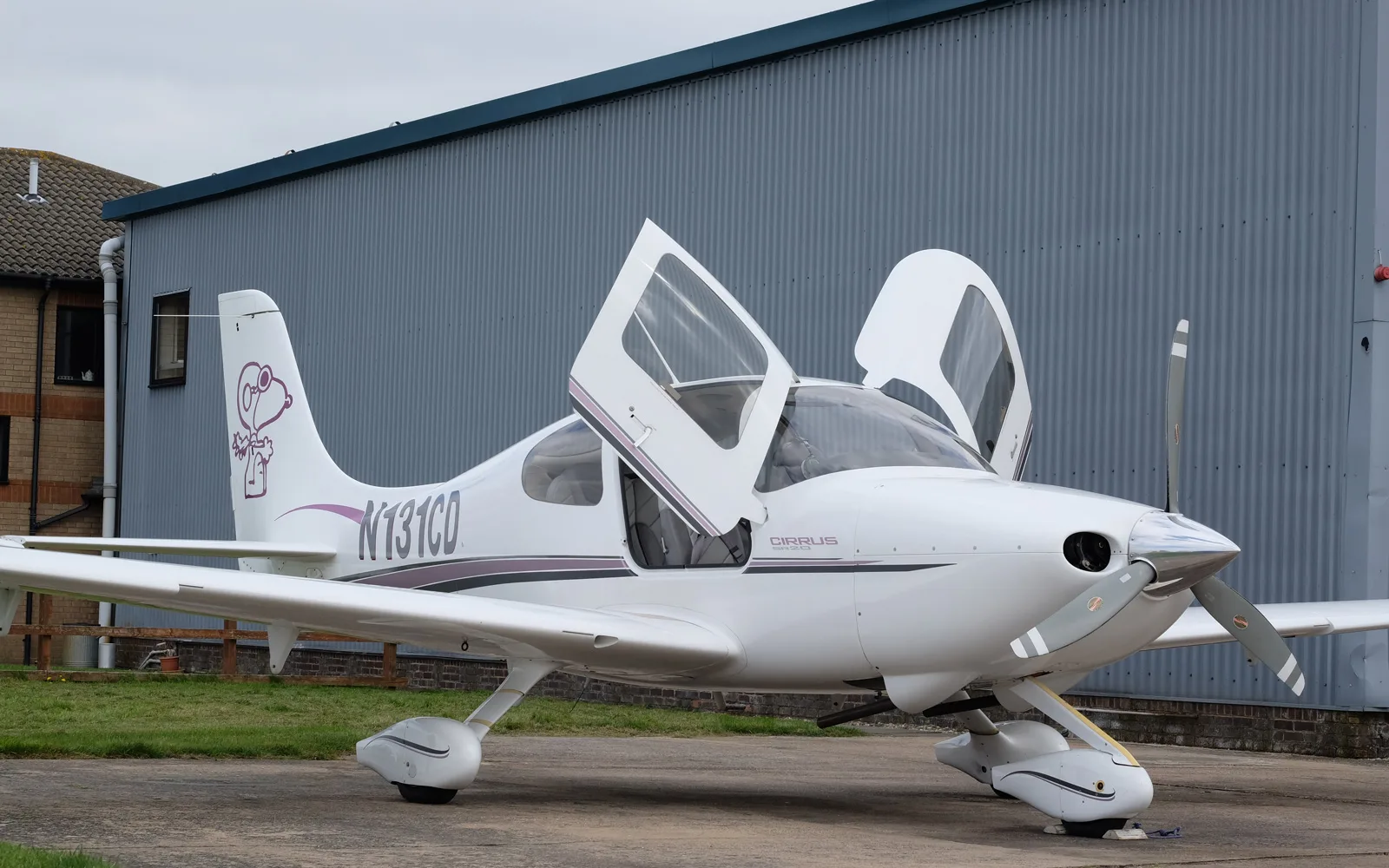 A white Cirrus SR22 aircraft with open gull-wing doors and a Snoopy graphic on the tail, parked next to a grey hangar.