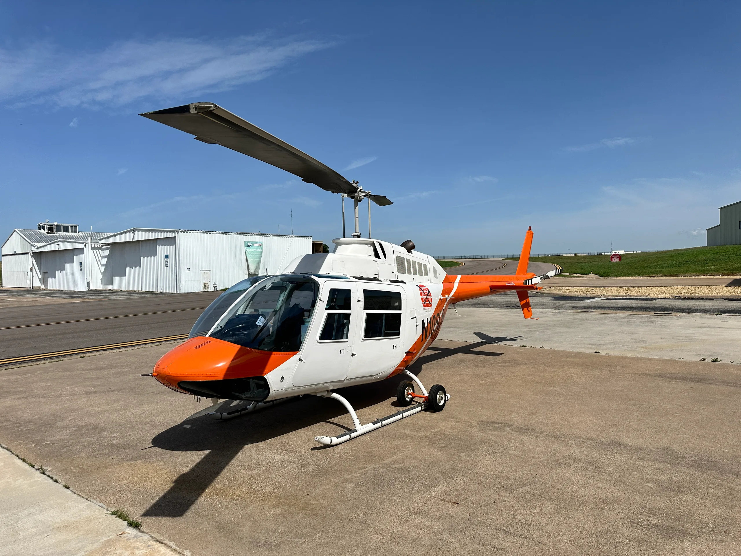 A white and orange helicopter, N1054, parked on an airfield with hangars under a blue sky.