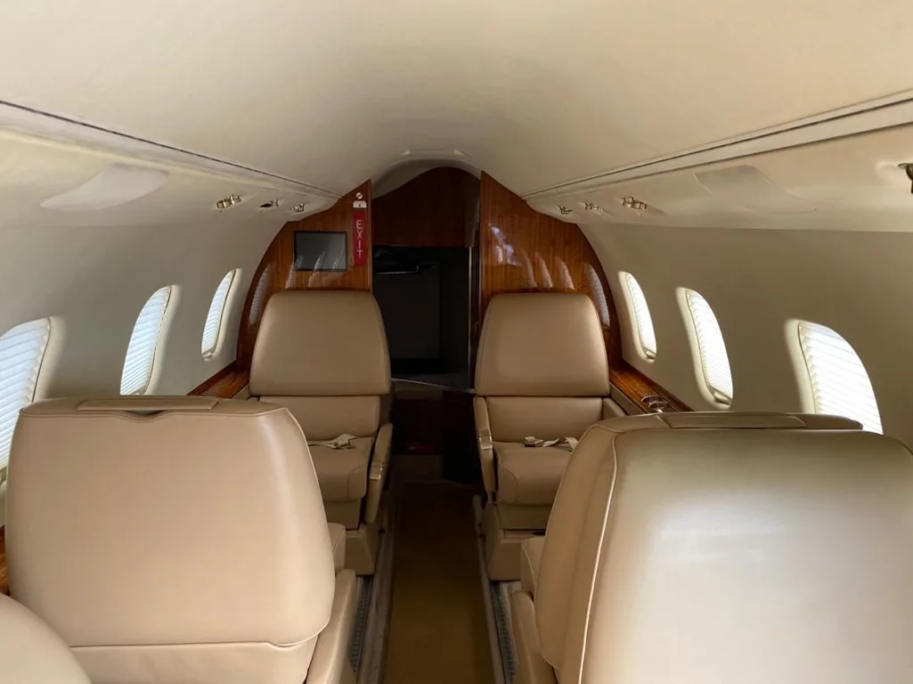 A view down the aisle of a private jet's cabin, showing beige leather seating and rich wood cabinetry.