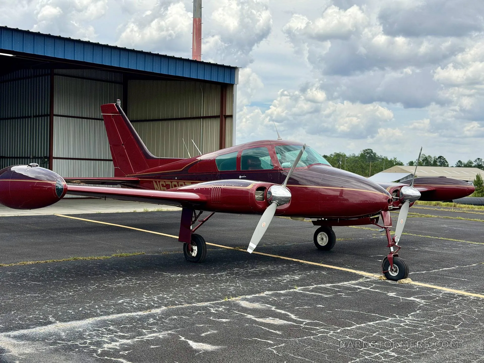 A red twin-engine aircraft, likely a Beechcraft Baron, with registration N6792T, parked on an asphalt apron in front of a blue hangar.