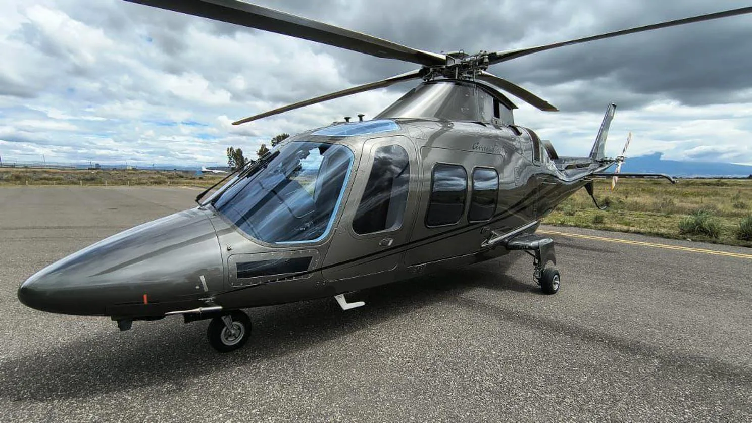 A sleek grey AgustaWestland AW109 GrandNew helicopter is parked on an asphalt tarmac under a cloudy sky.