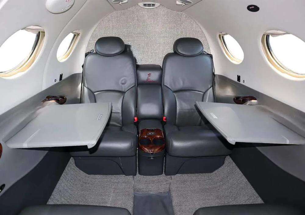 View of the aircraft's cabin interior showing two dark gray leather seats with fold-out tables and a center console.