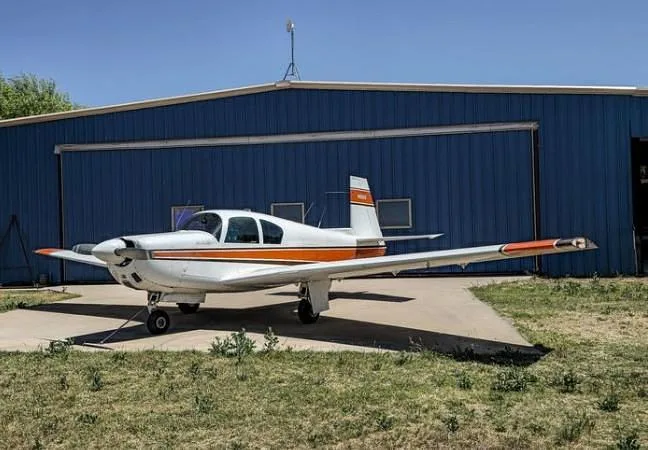 A white and orange Mooney M20 aircraft parked on concrete in front of a blue hangar, viewed from the side.
