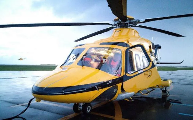 A yellow helicopter with black accents sits on a wet tarmac, with another helicopter in the distance.