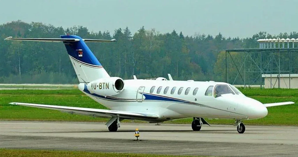 A white business jet with blue and gold stripes, registration YU-BTN, parked on an airfield with trees in the background.