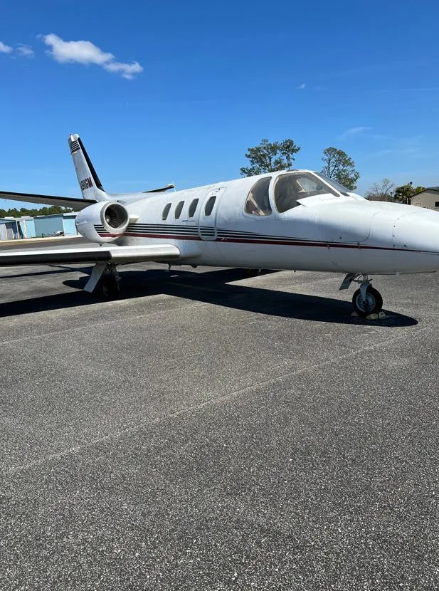 A white Cessna Citation I business jet with red and black stripes is parked on an asphalt tarmac under a clear blue sky.