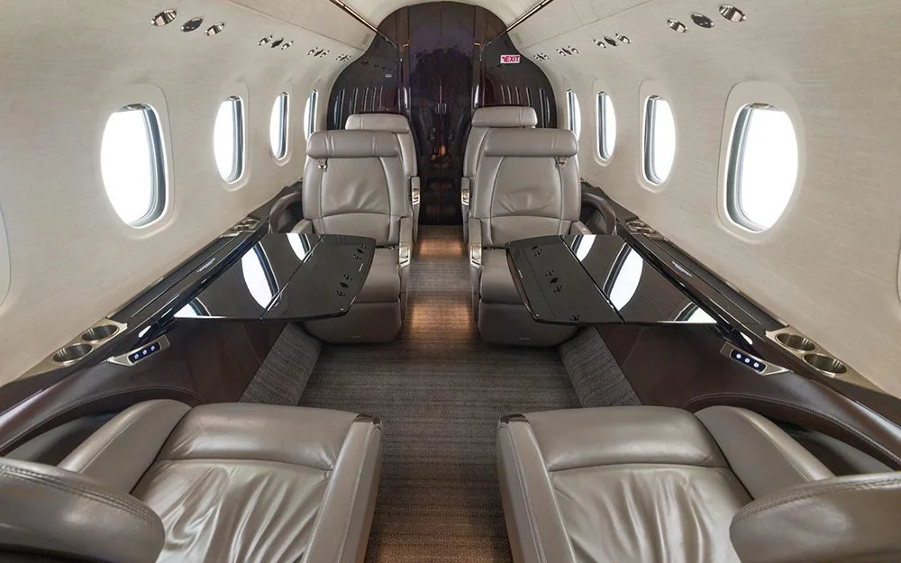 The interior cabin of a private jet, showing light brown leather seats with black fold-out tables deployed.
