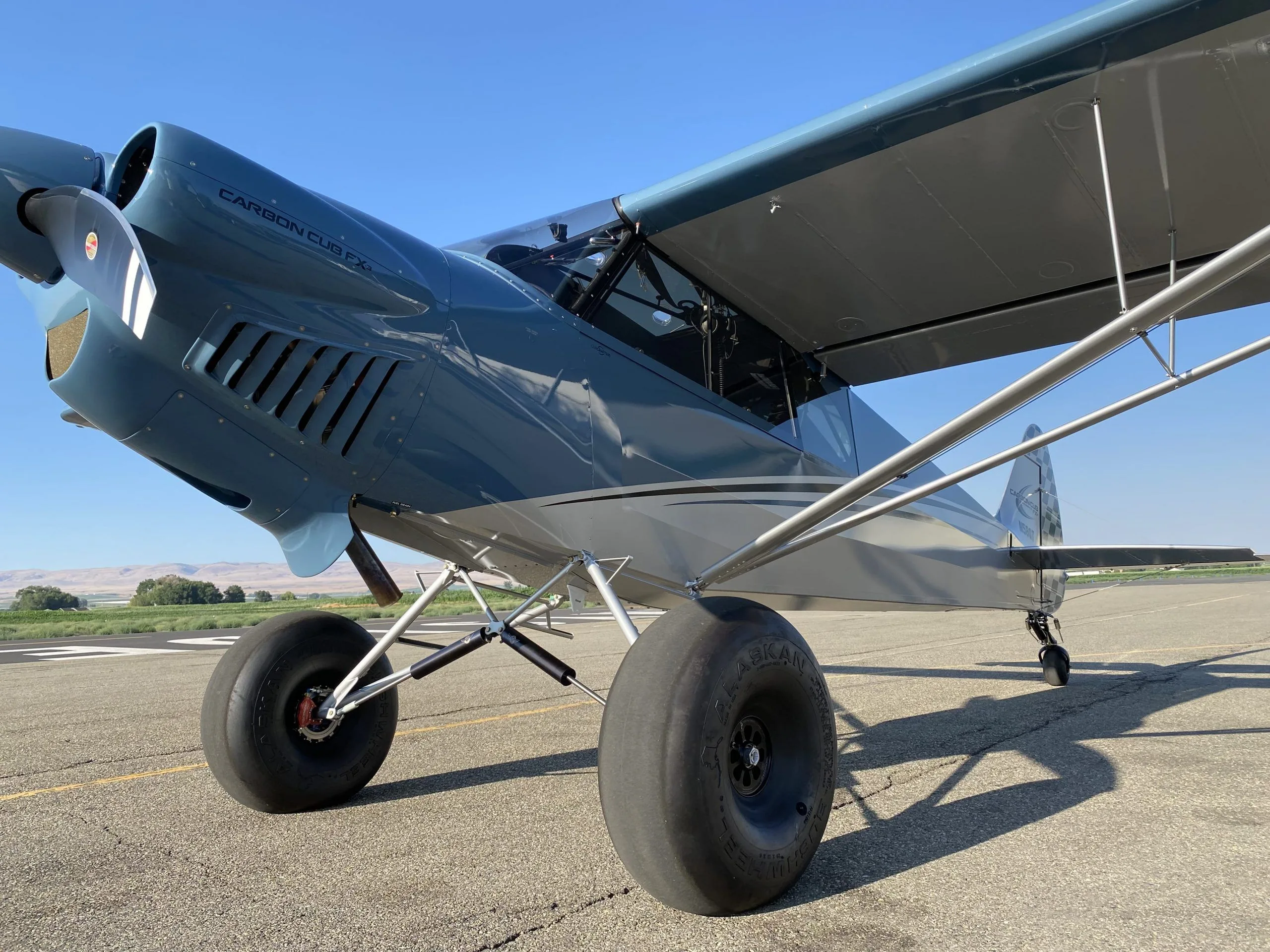 A close-up view of the front and side of a blue and grey Carbon Cub FX-3 aircraft, highlighting its large tundra tires.