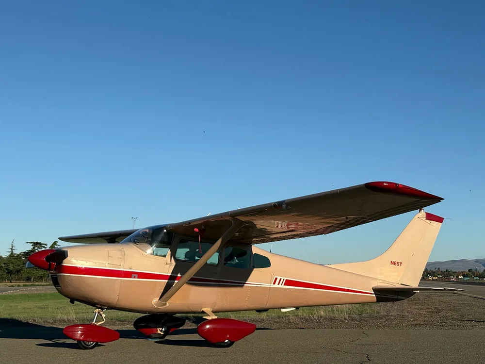 A beige and red Cessna 182 aircraft, registration N85T, parked on an airfield under a clear blue sky.