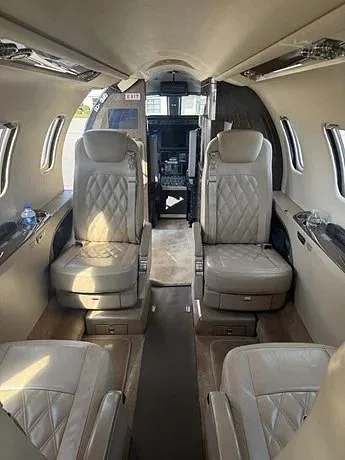 A view down the aisle of a private jet's cabin, showing light tan leather seats with diamond stitching and the cockpit entrance in the background.