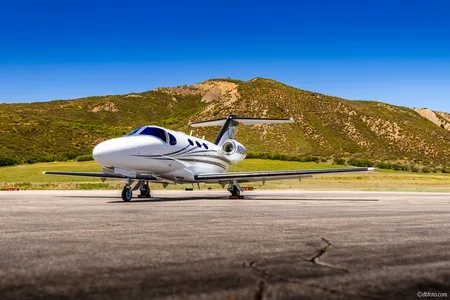 A white business jet with dark and light grey stripes is parked on an asphalt runway with green, treeless mountains under a clear blue sky in the background.