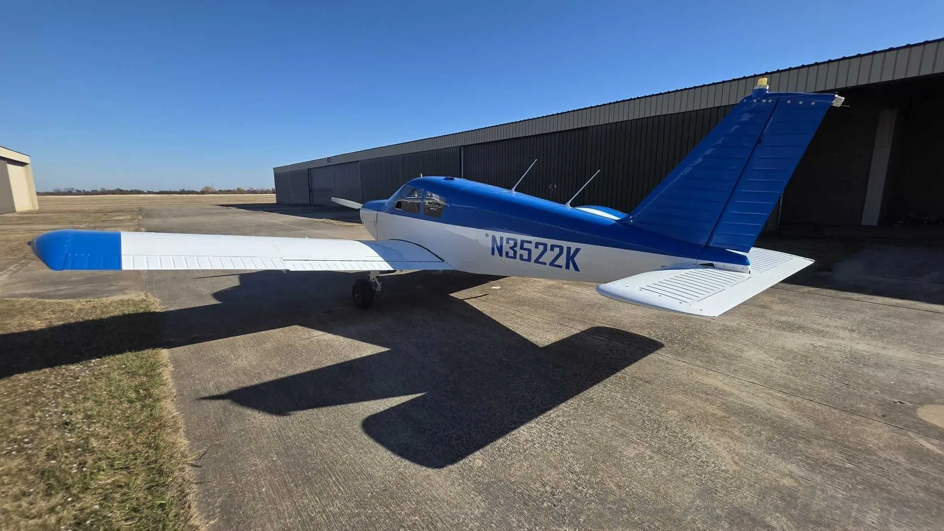 A blue and white single-engine aircraft, N3522K, parked on an asphalt ramp next to a hangar.