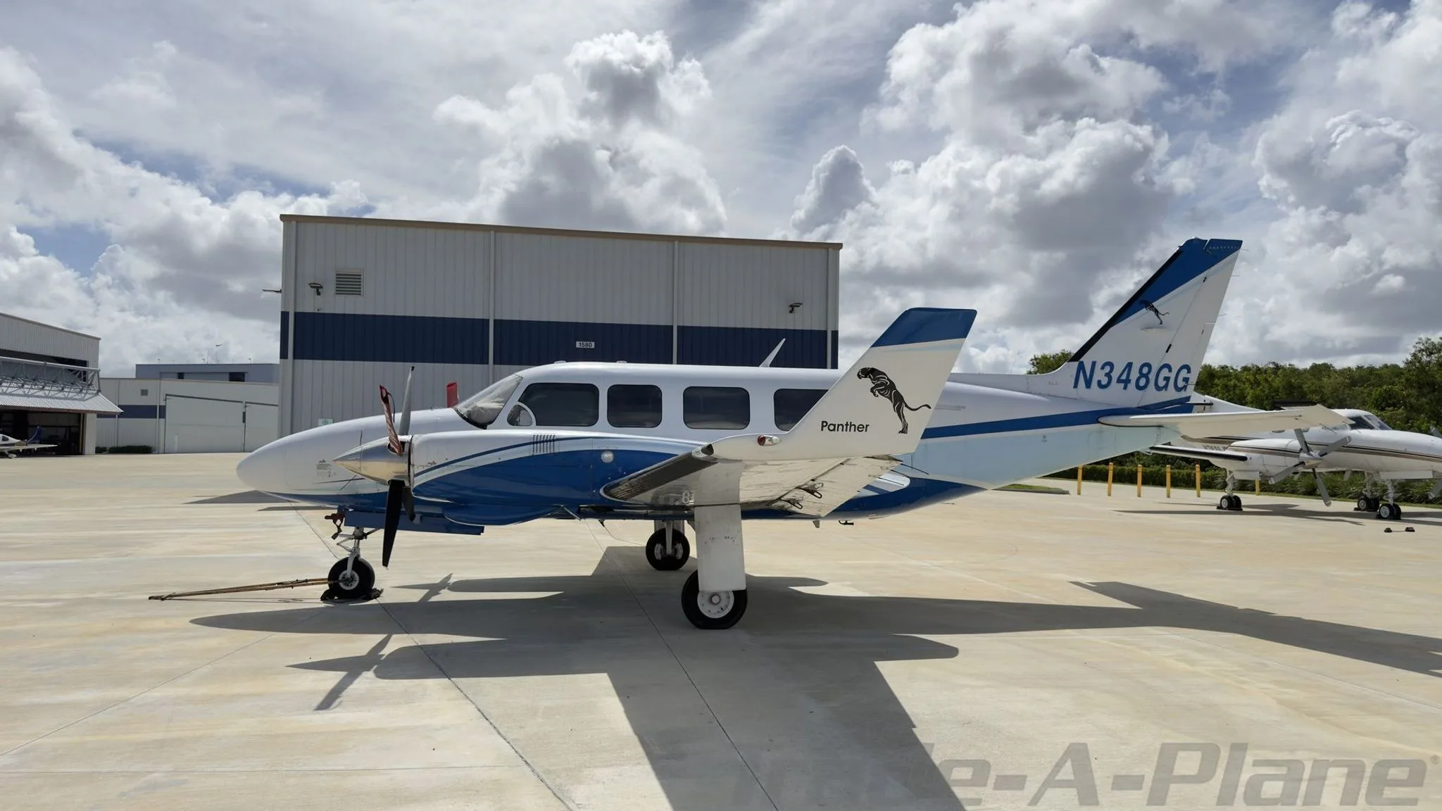 A white and blue twin-engine propeller aircraft, N348GG, with a 'Panther' decal, parked on a concrete ramp in front of hangars.